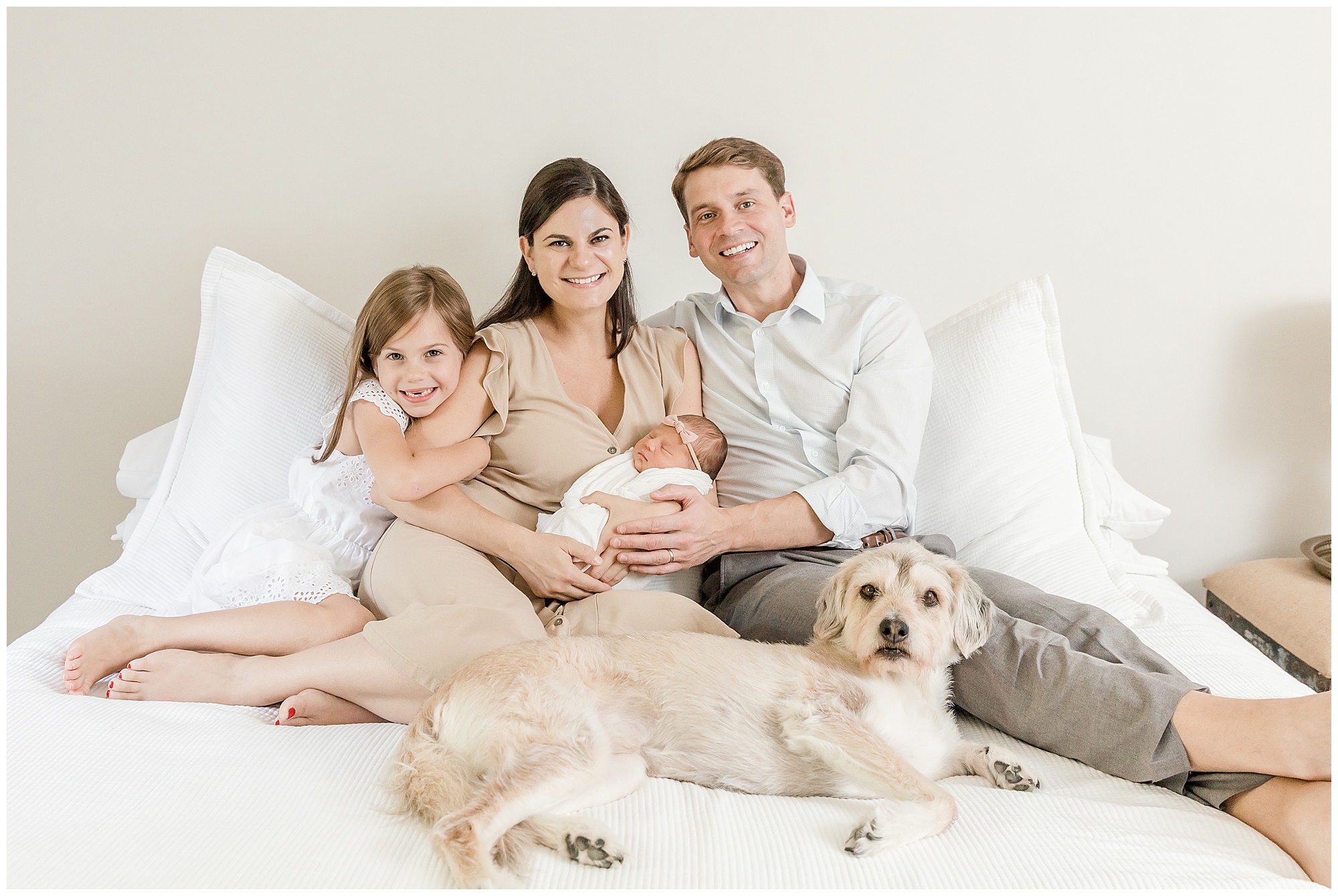 Family portrait of parents with their two daughters and dog sitting on a white bed.