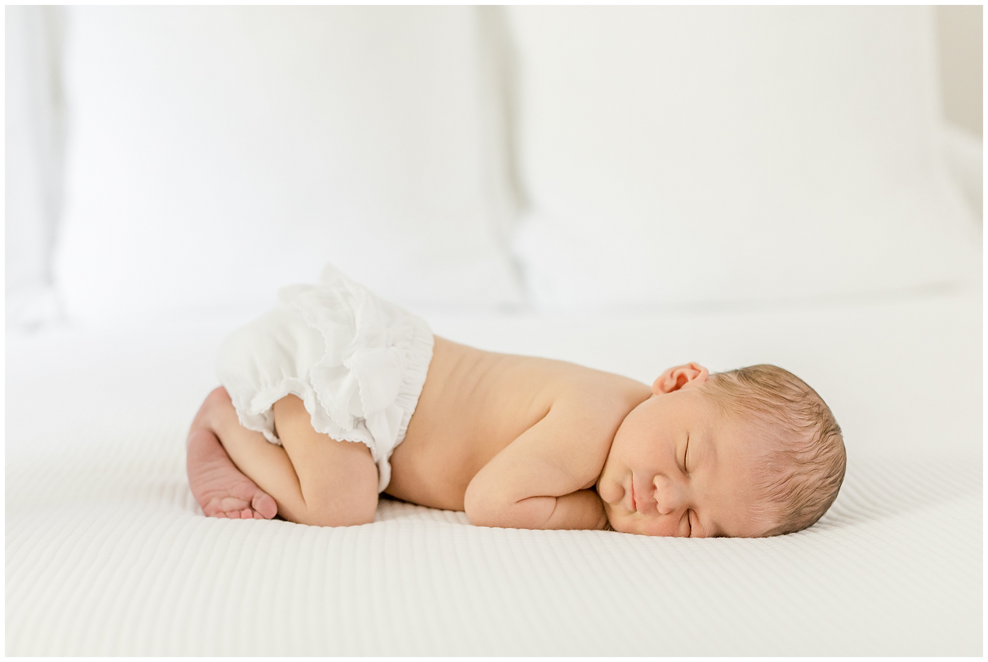 Photo of a newborn baby girl in bloomers posed on a white comforter.