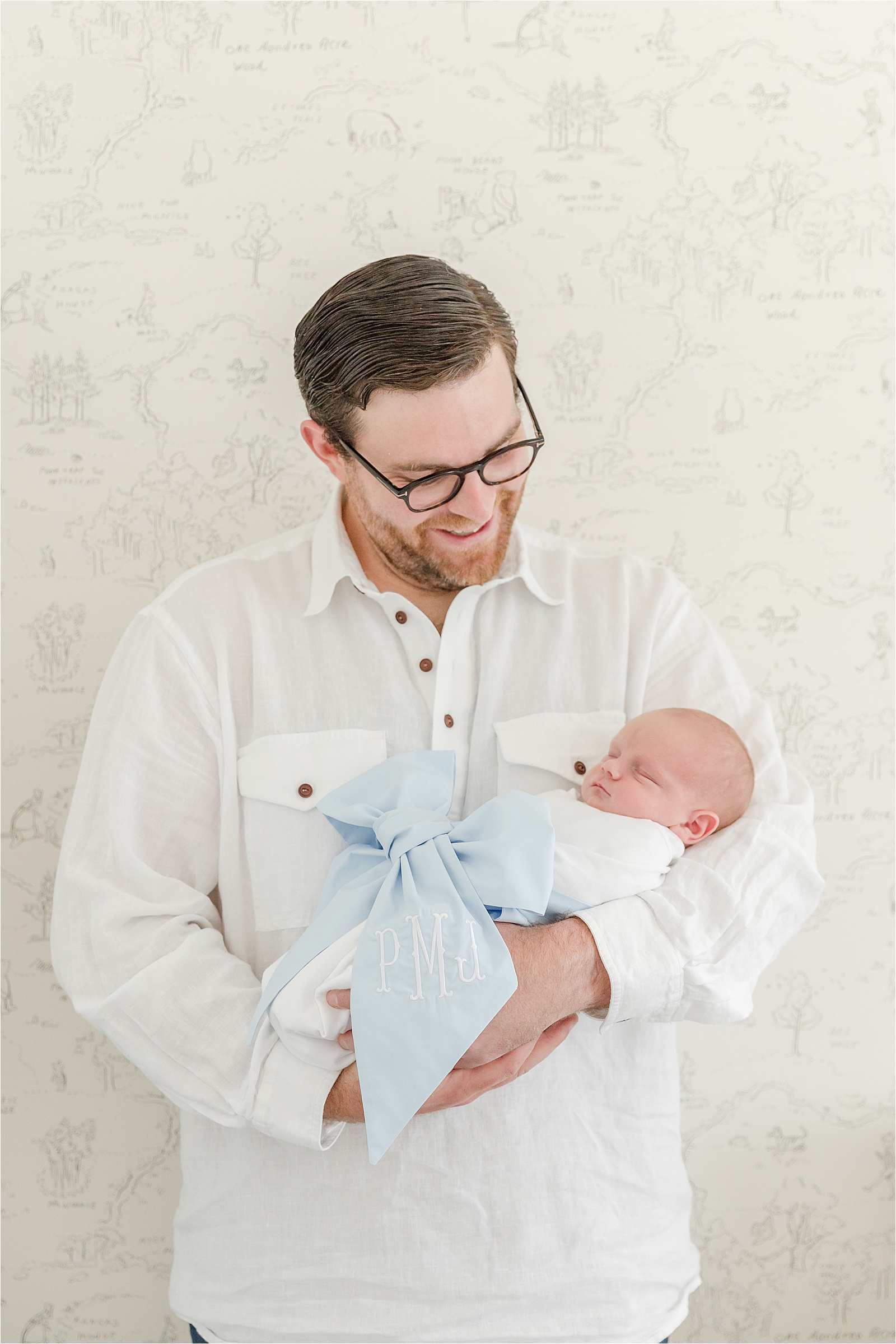 Father holding newborn son in Beaufort Bonnet Bow Swaddle in front of Hundred Acre Wood wallpaper