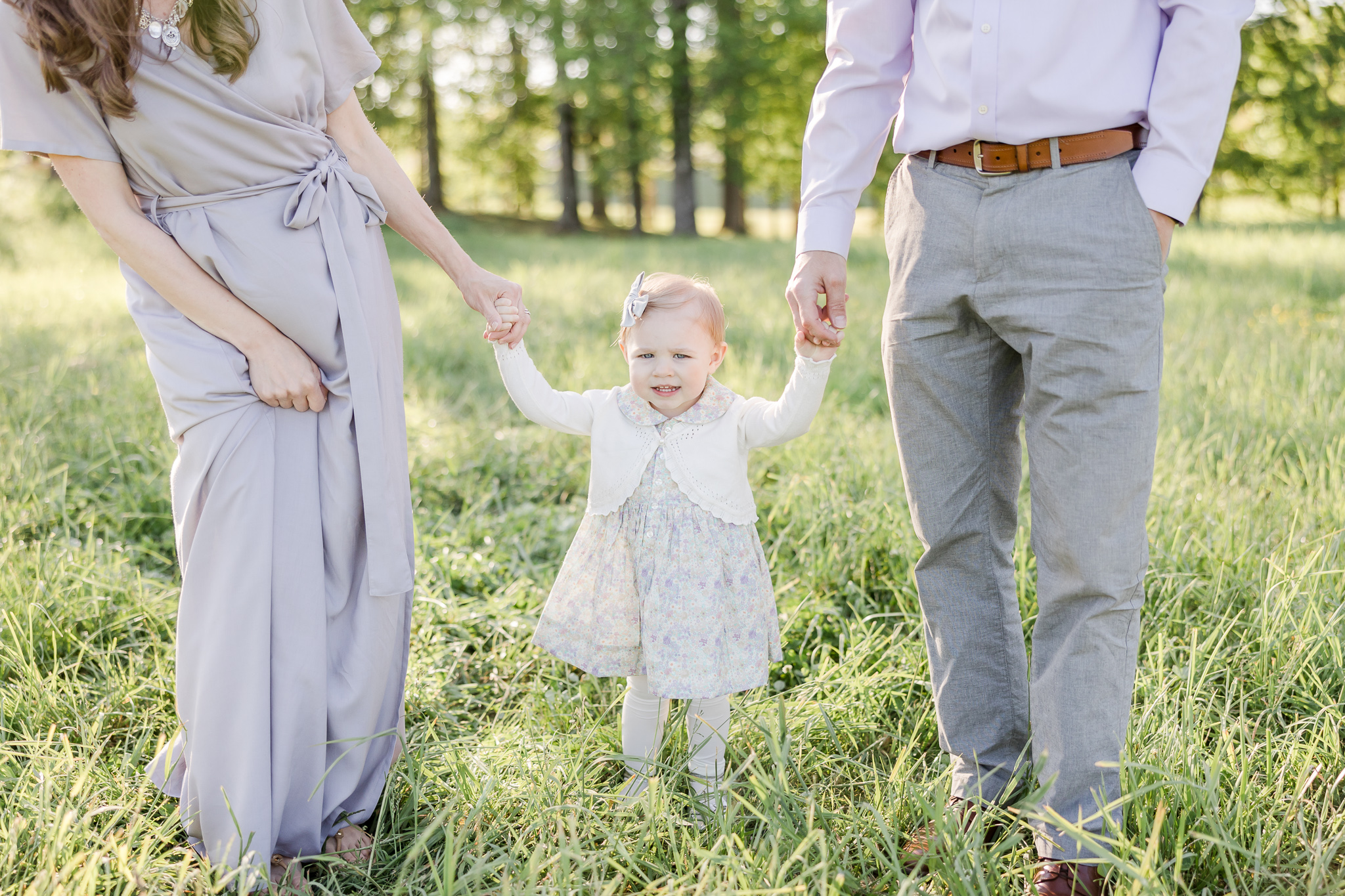 Two parents holding the hand of their toddler daughter in a grassy field.