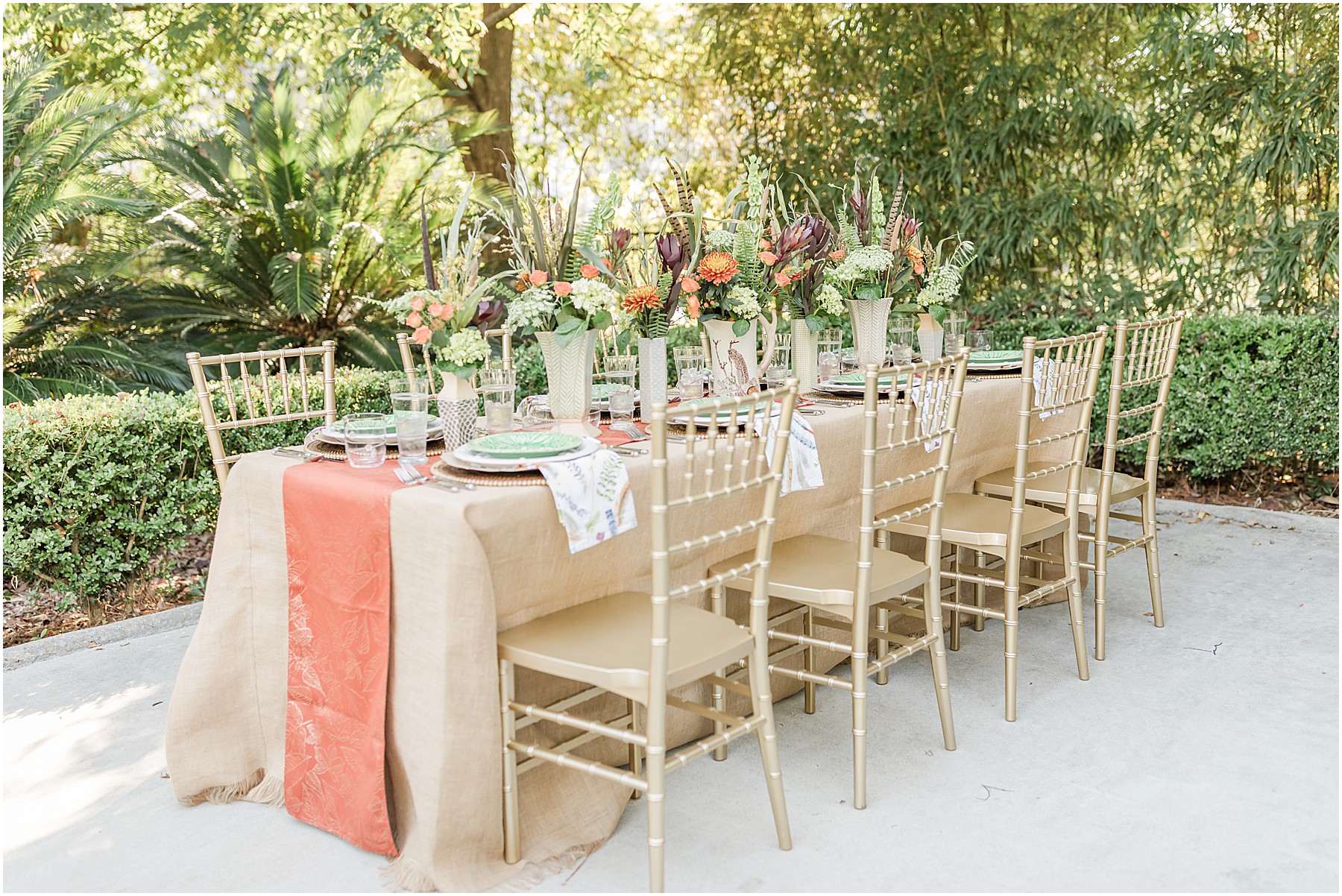 A long Fall table covered with a burlap tablecloth and orange runner, surrounded by eight bamboo golden chairs and covered in an eight person places setting with a row of vases down the center with fall flowers.