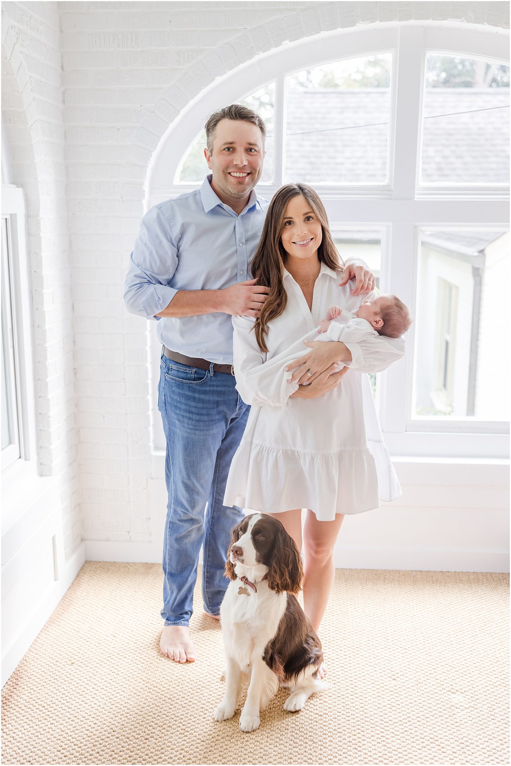 New mother and father posing with their newborn and dog in front of a large window for their Greenville in home newborn photos.
