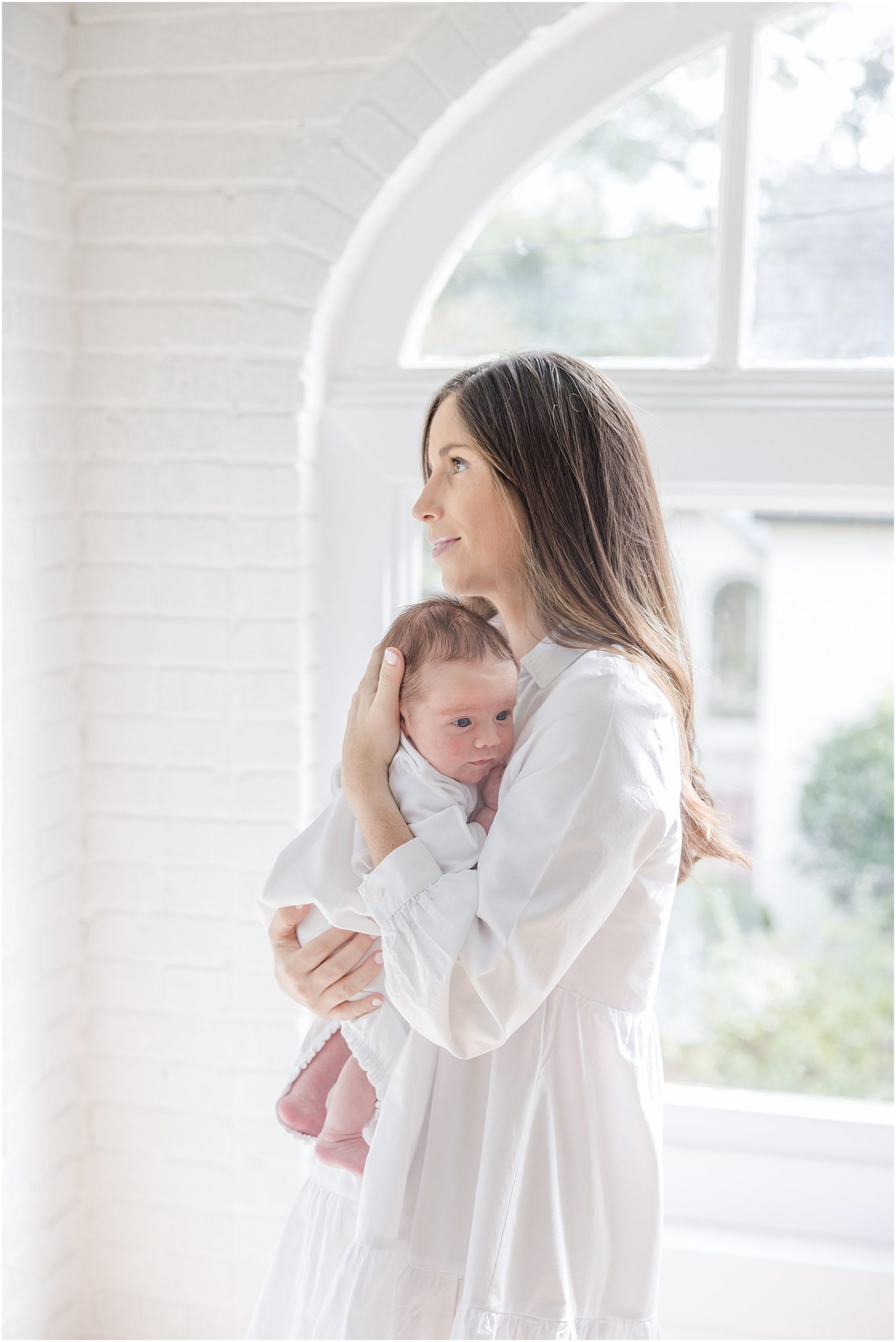 Young mother gazing out the window while cradling her newborn baby in this Greenville in home newborn photography session.
