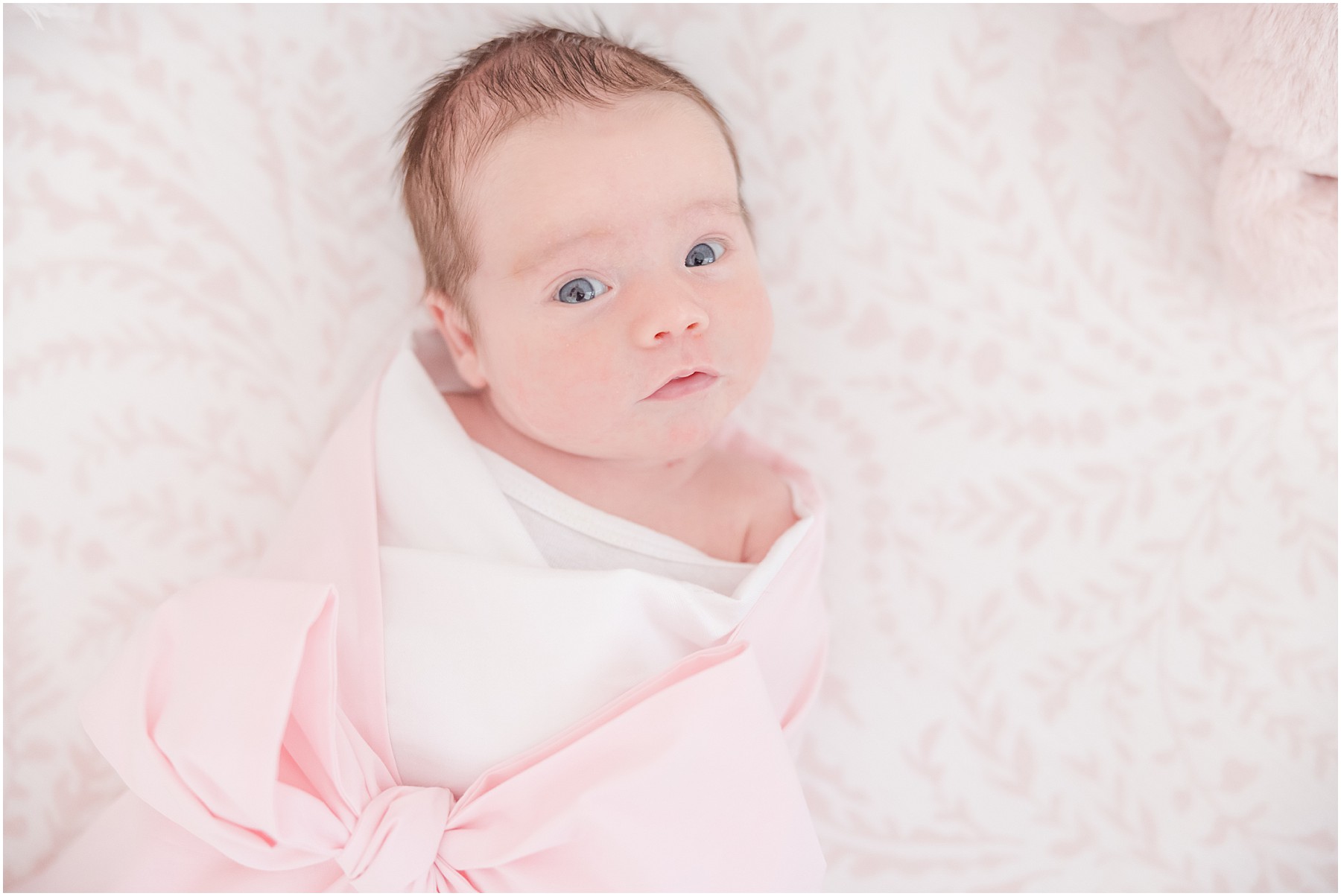 A baby in a pink bow swaddle gazes toward the camera.