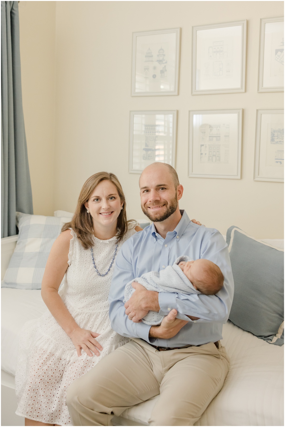 Greenville Newborn Photography of parents seated on a white daybed holding a newborn boy wrapped in a blue blanket.