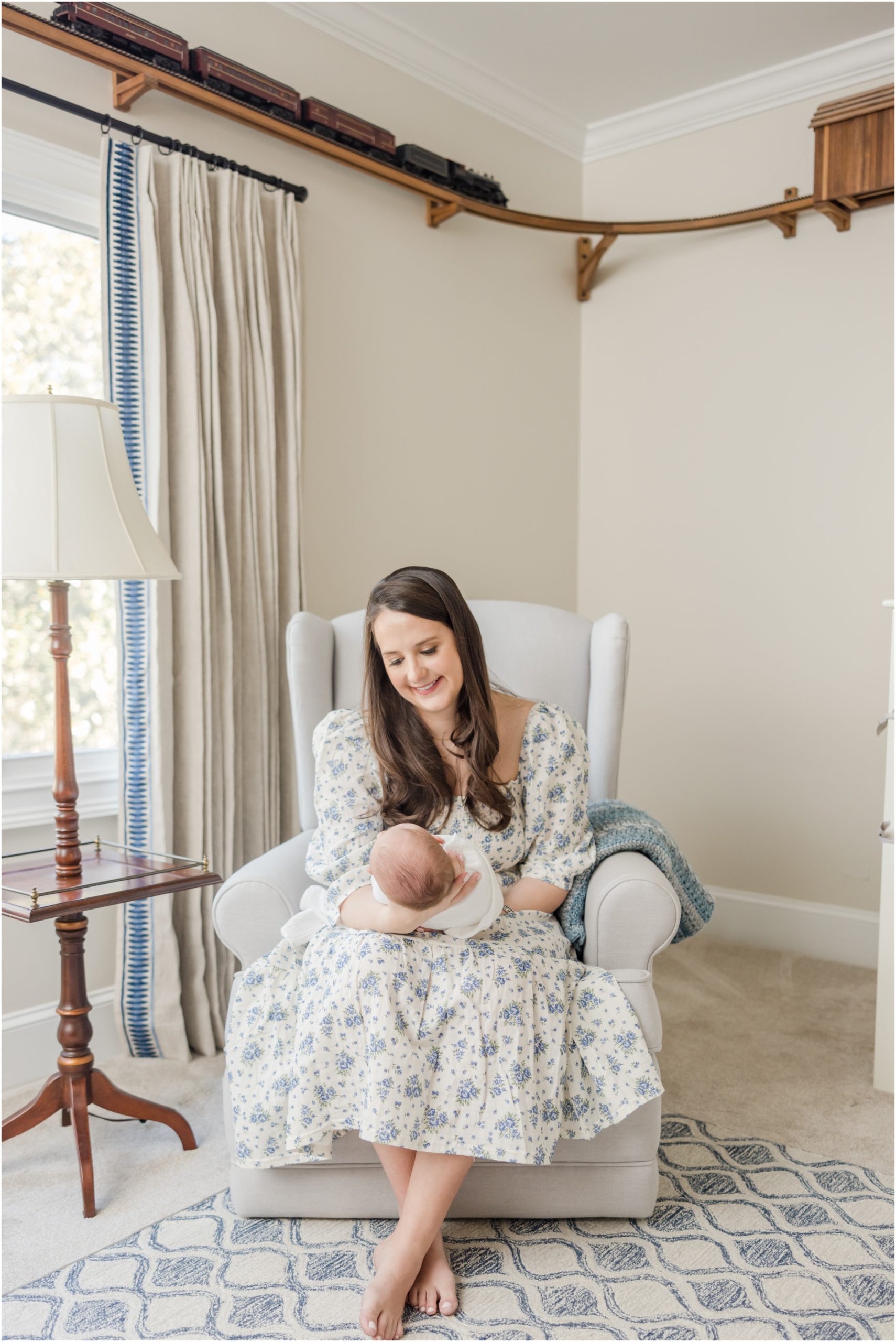 A mother is seated in a nursery glider and smiling down at her newborn son in her hands.