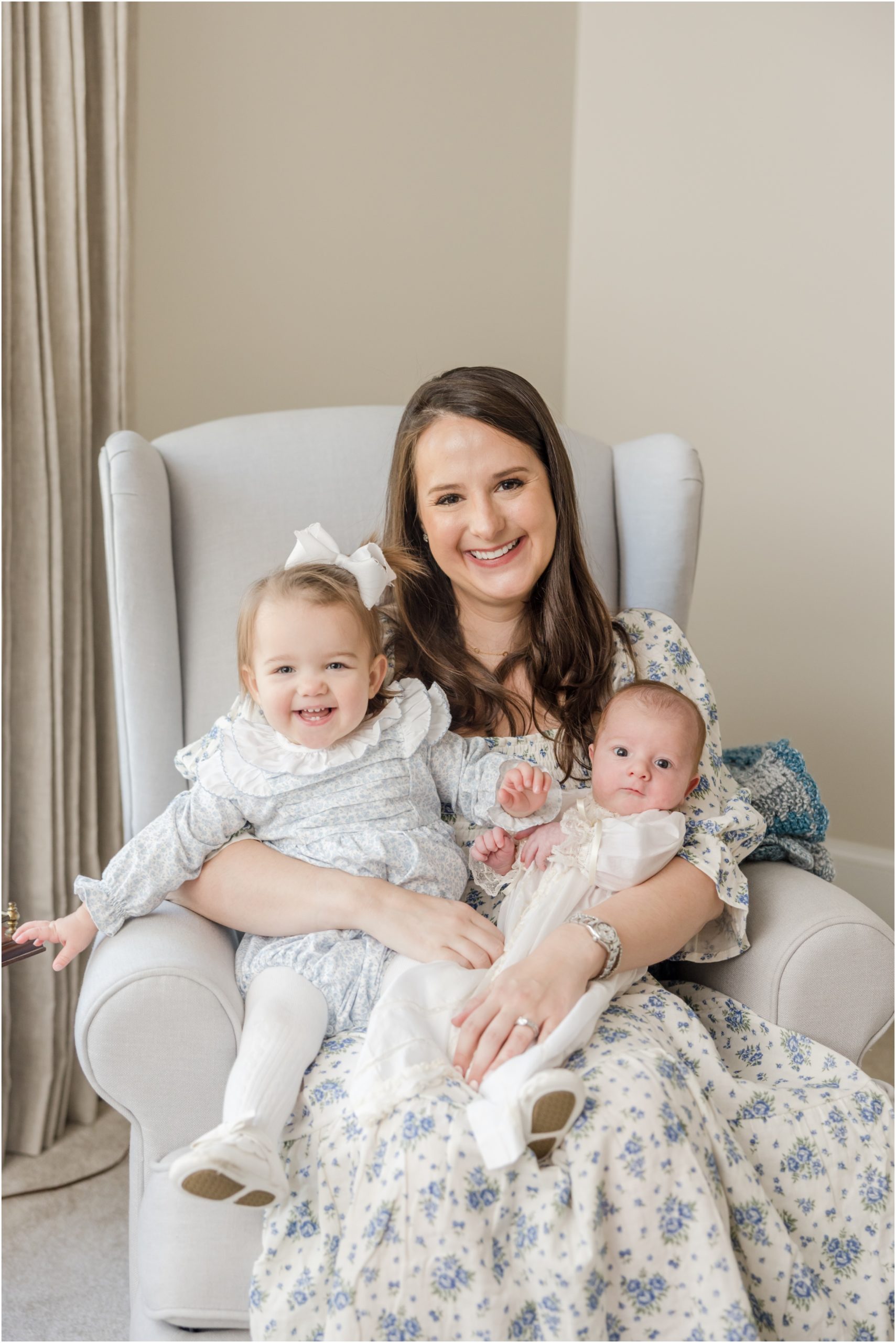 Portrait of a mother in a blue floral dress seating in a glider holding her toddler daughter and newborn son during her Greenville newborn photography session.