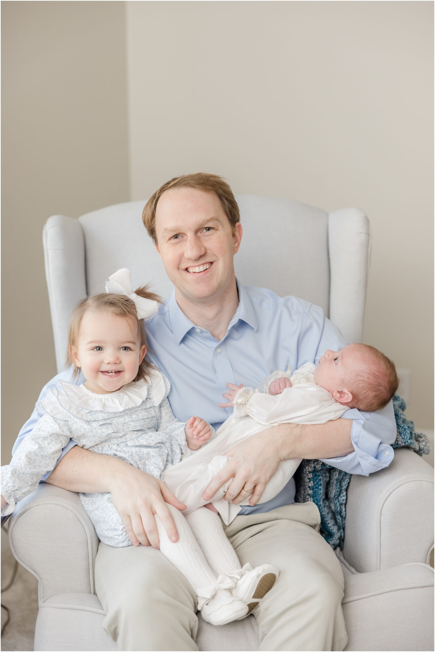A father seated in a rocker holding his toddler daughter and newborn son during a Greenville newborn photography sesion.