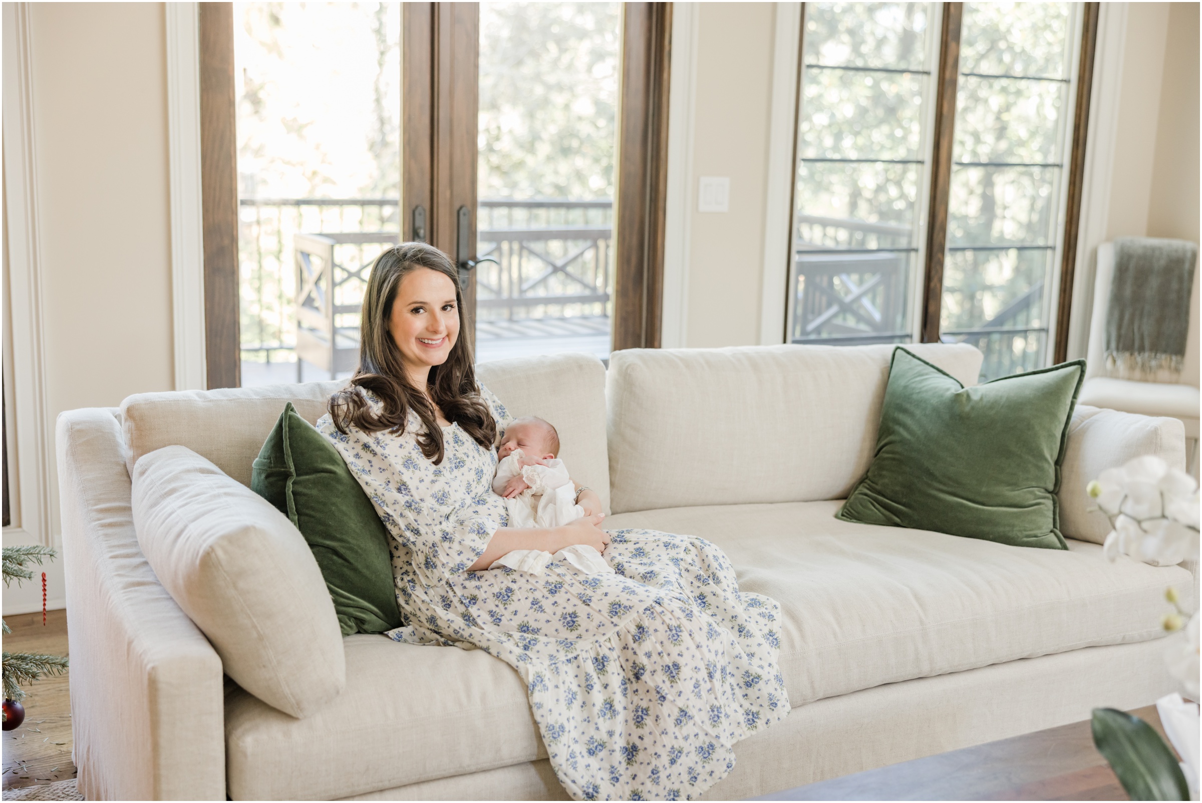 Portrait of a young mother in a blue floral dress holding her newborn son on a tan couch in front of large windows smiling for a photo taken by a Greenville newborn photographer.