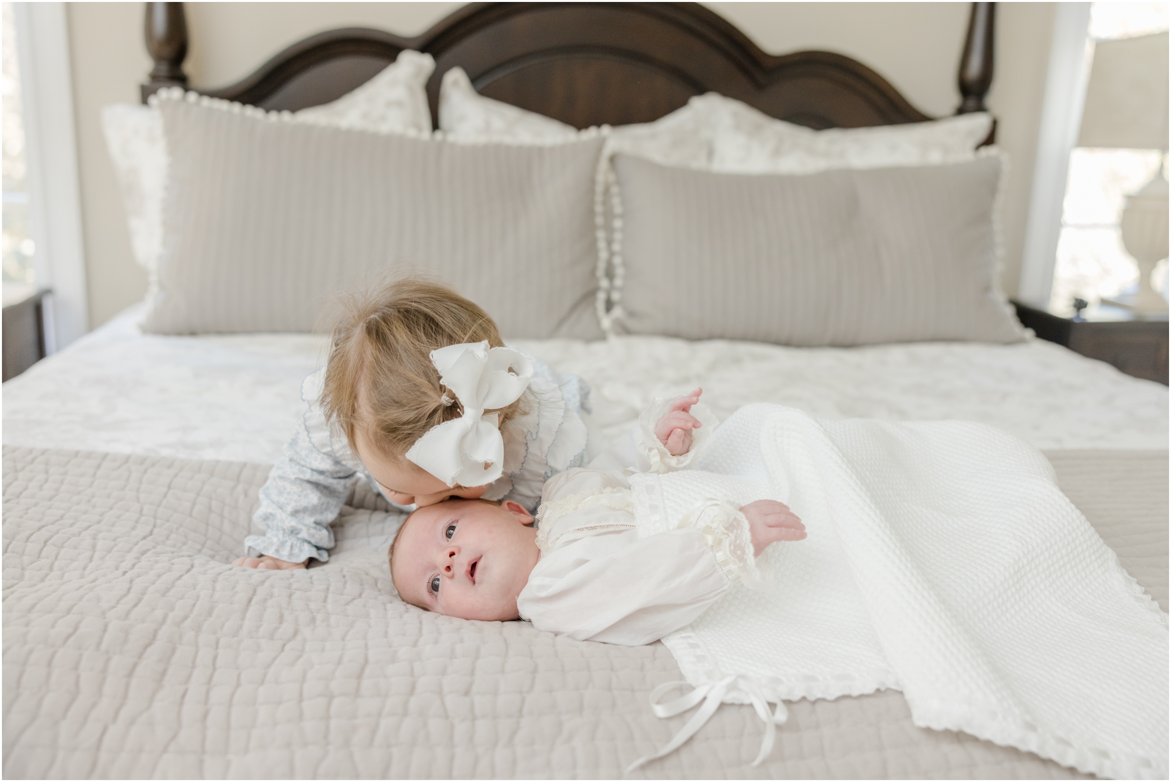 A toddler girl leans over to kiss the head of her newborn baby brother who is laying on their parent's bed during a Greenville newborn photography session.