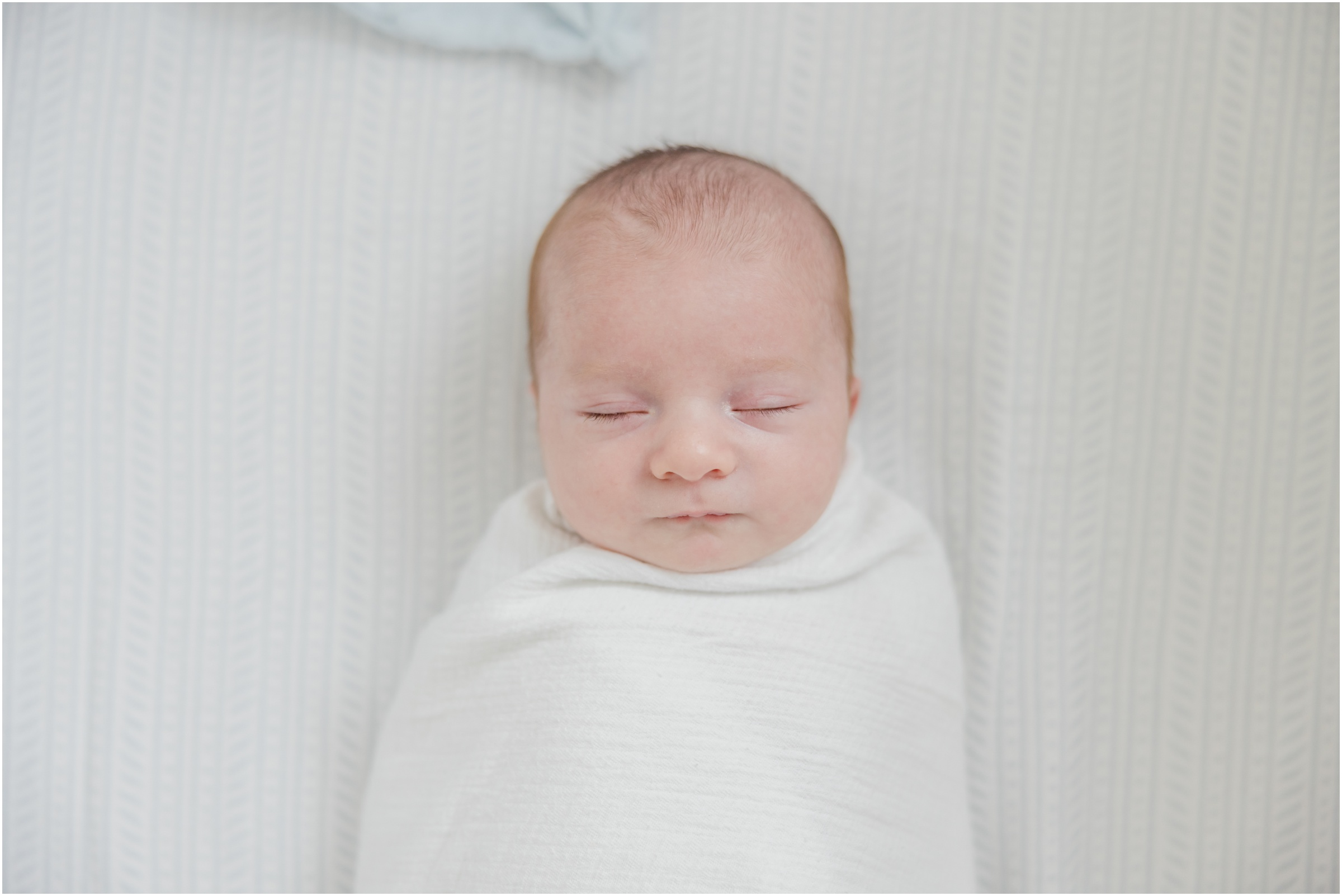 Portrait of a newborn baby boy swaddled in a white blanket laying on a soft blue patterned crib sheet.