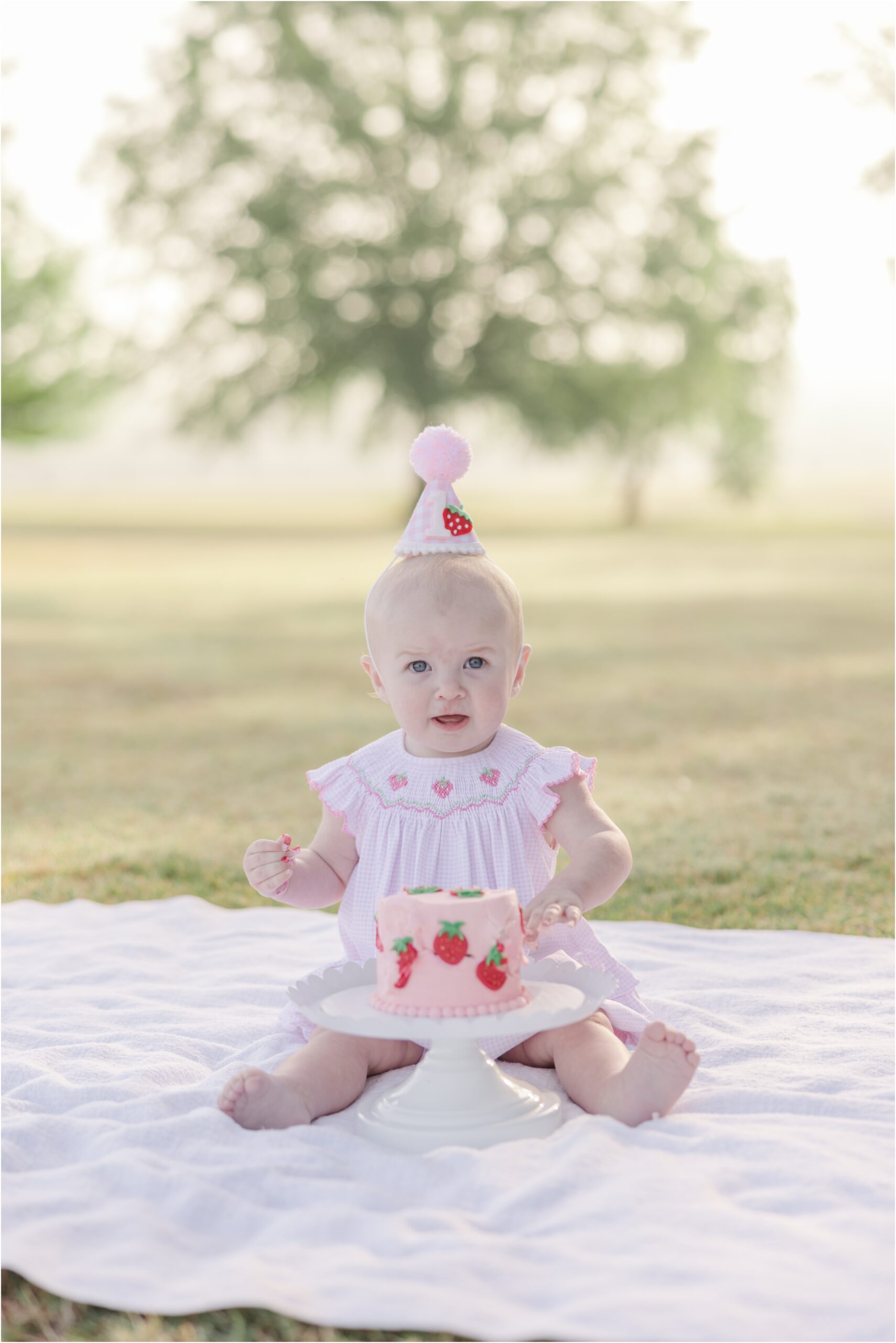 First birthday portraits of a baby girl with a strawberry smash cake, strawberry bubble, and birthday hat with a one and strawberry on it.