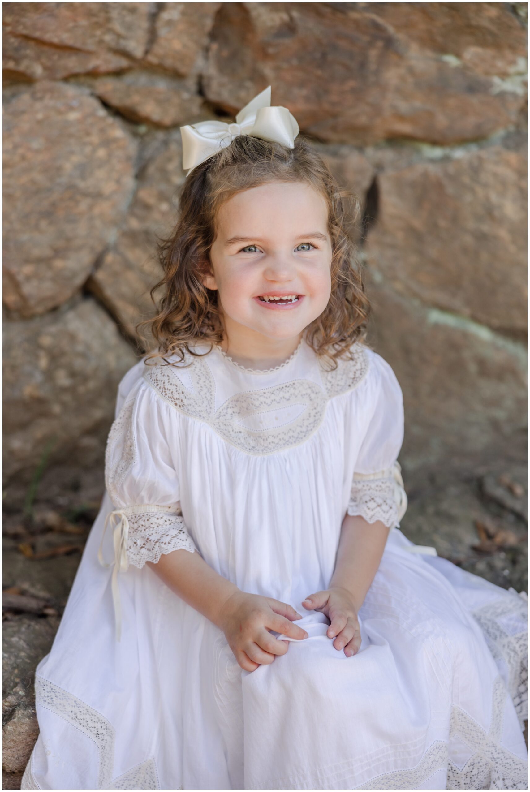 Young girl posing for Greenville children's portraits in a white dress with lace detailing in front of a rock wall backdrop.