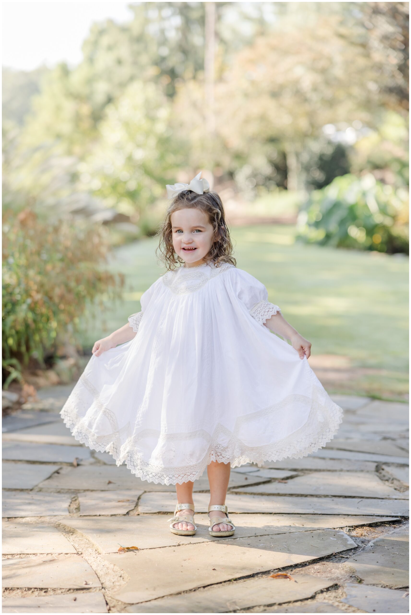 Two year old girl standing in a garden holding out a white heirloom dress for a photo taken by Greenville children's photographer Molly Hensley.