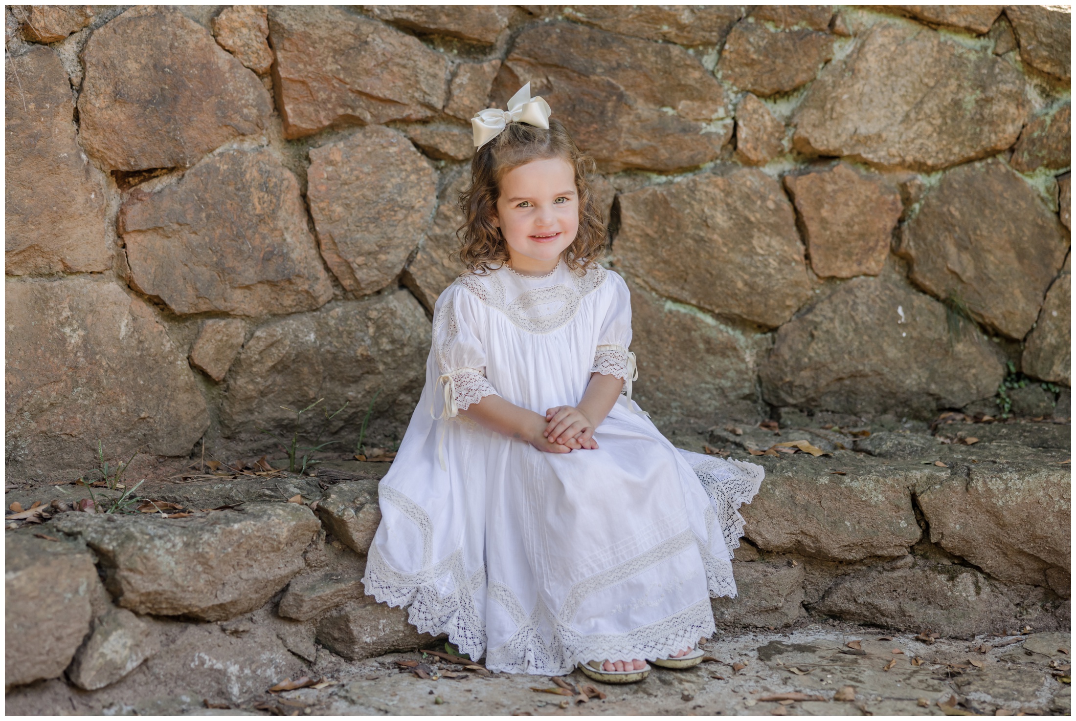 Greenville children's portraits of a young girl wearing a white heirloom lace dress sitting on a rock wall bench.