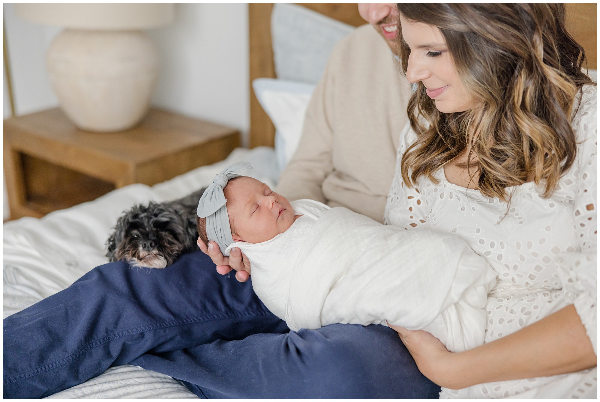 Parents and a small black dog sit on their bed gazing down at their newborn baby girl taken by Greenville SC in. home newborn photographer.