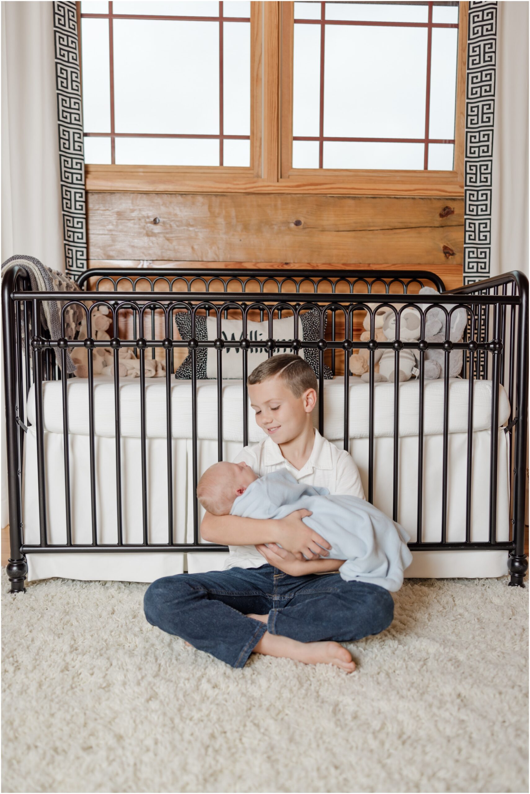 A young boy sits on a shag rug holding his newborn baby brother in front of a black and white cabin nursery during a Greenville SC newborn photography session.