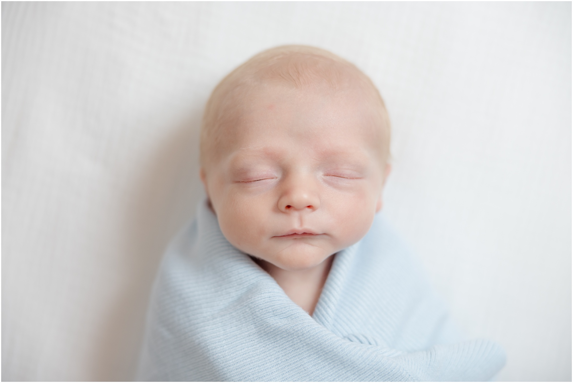 Portrait of a baby boy wrapped in a blue blanket on a white crib sheet during a Greenville SC newborn photography sesison.