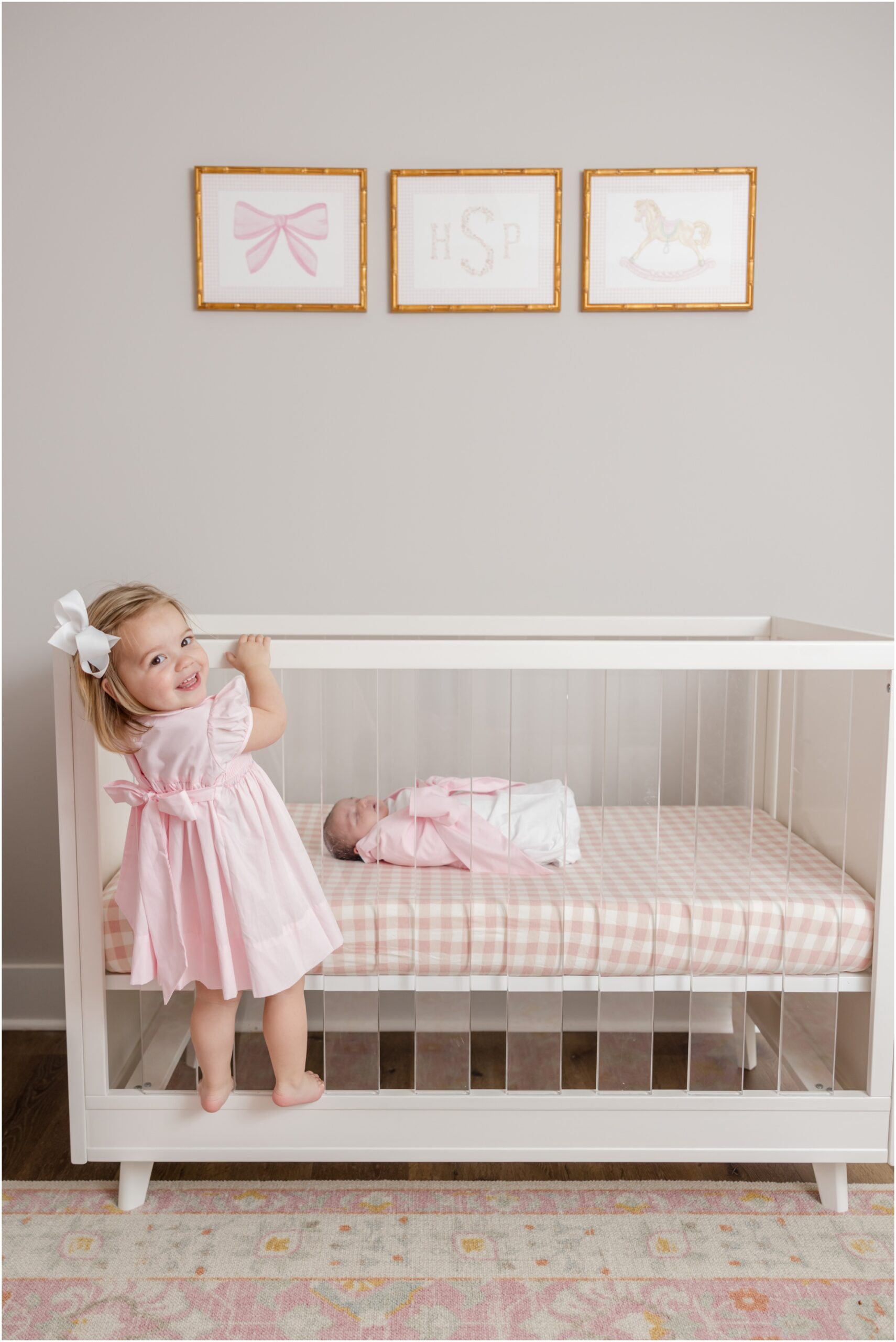A toddler girl wearing a soft pink dress stands on her newborn sisters crib and smiles over her shoulder at the camera during newborn photos in Greenville SC.