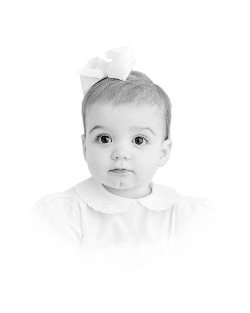 A black and white traditional heirloom portrait of a one year old girl with a serious expression in white with a white bow with a white vignette by Greenville photographer Molly Hensley.