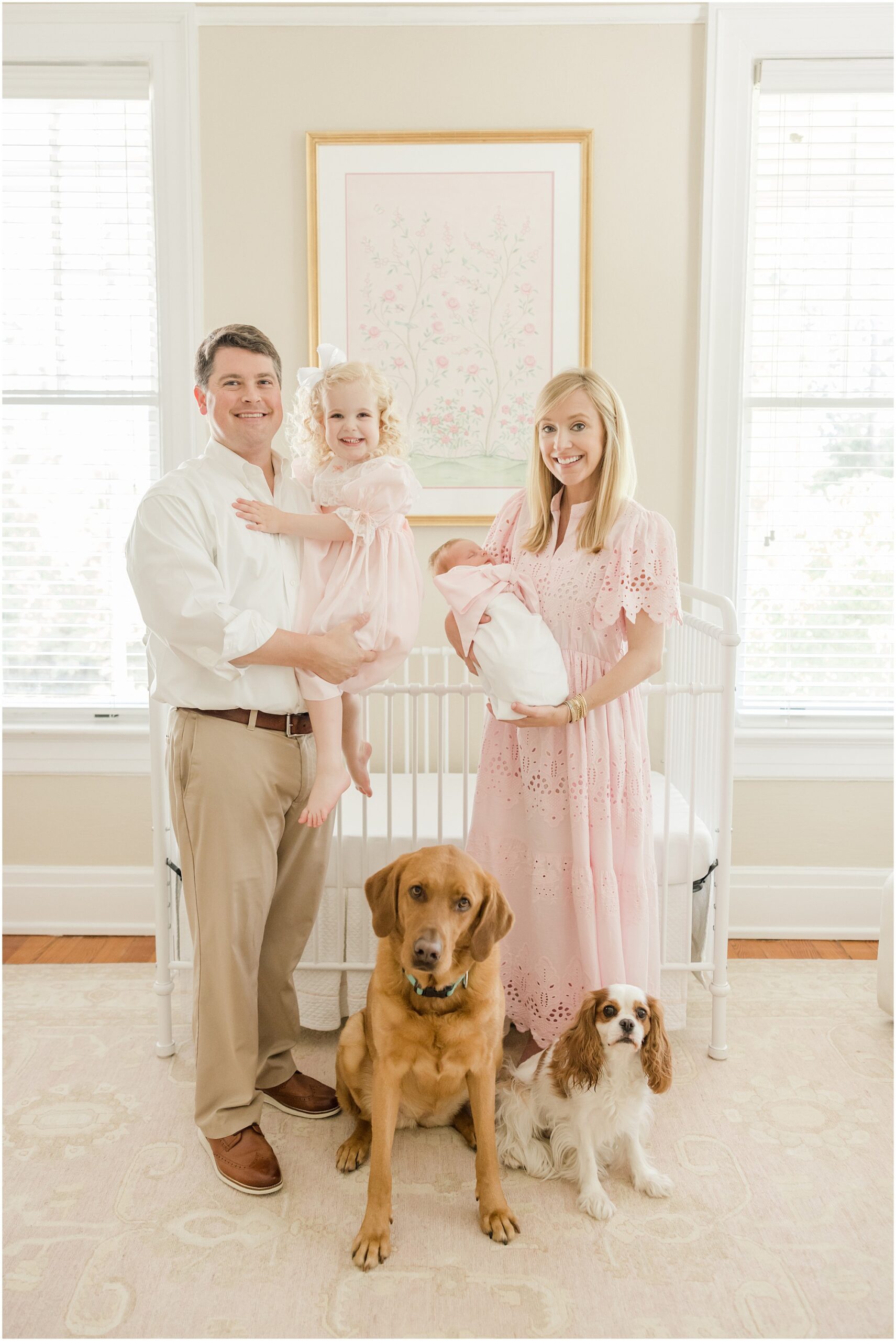 A family poses in front of a crib holding a toddler and newborn daughter while two dogs sit at their feet.