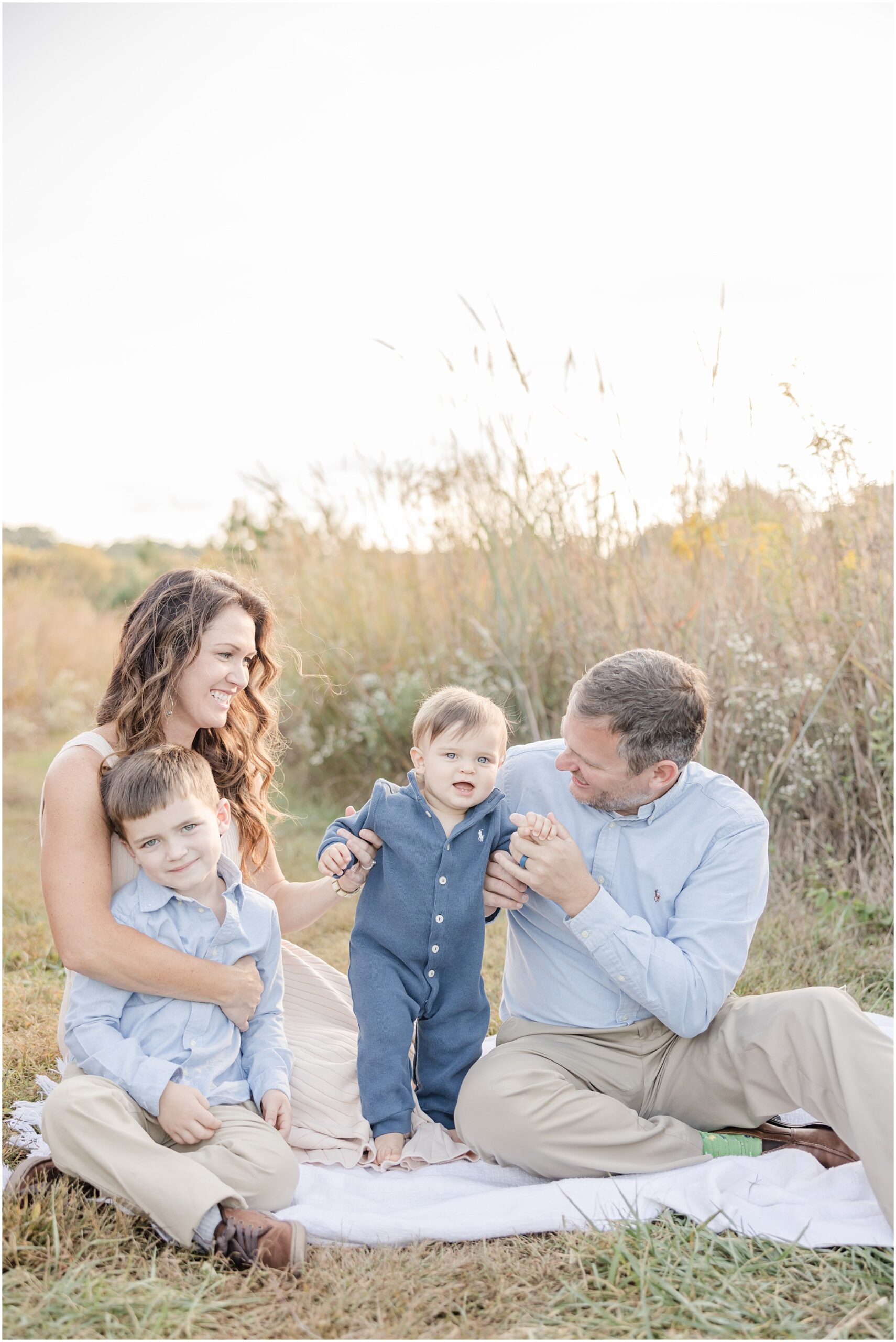 Parents sit on a white blanket in a field with their two young boys, smiling at one another while a Greenville family photographer takes their photo.